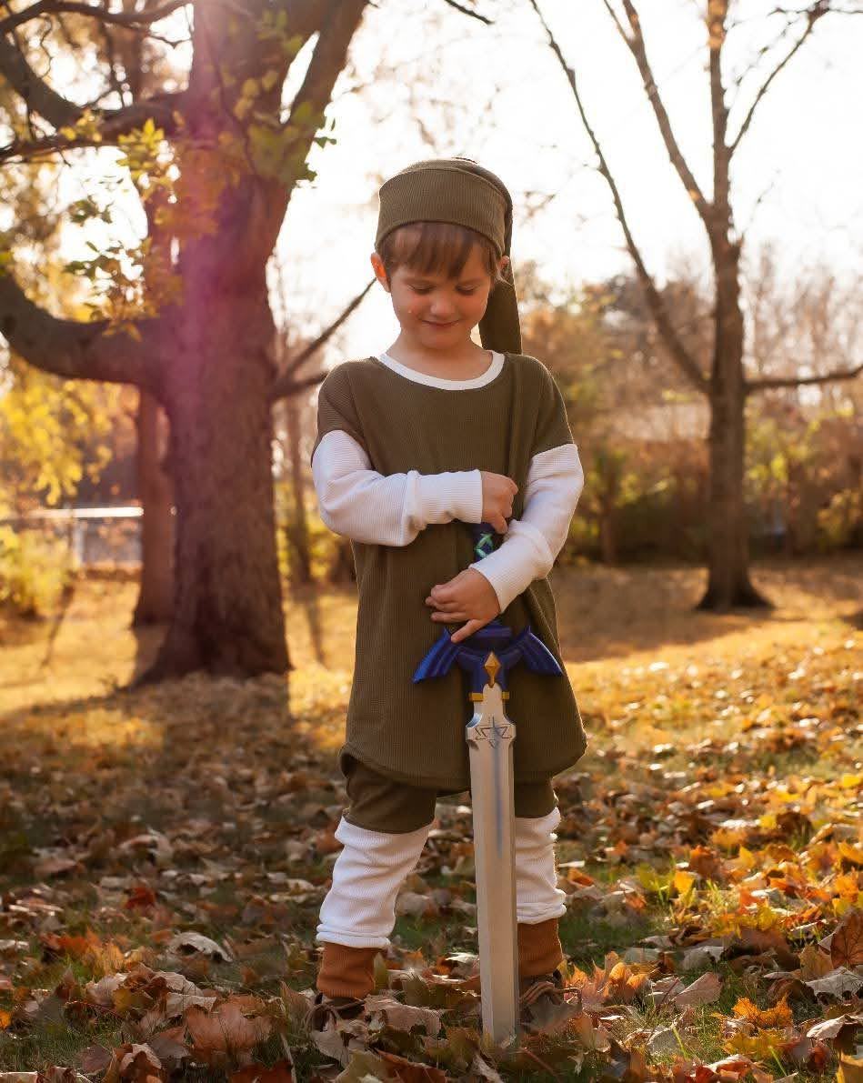 A young boy in a green costume tunic standing under bare trees