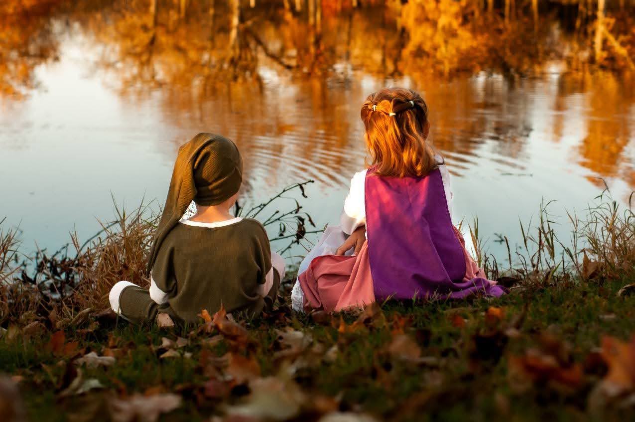 Two children in dress-up costumes sitting by a pond, autumn trees reflected in the water
