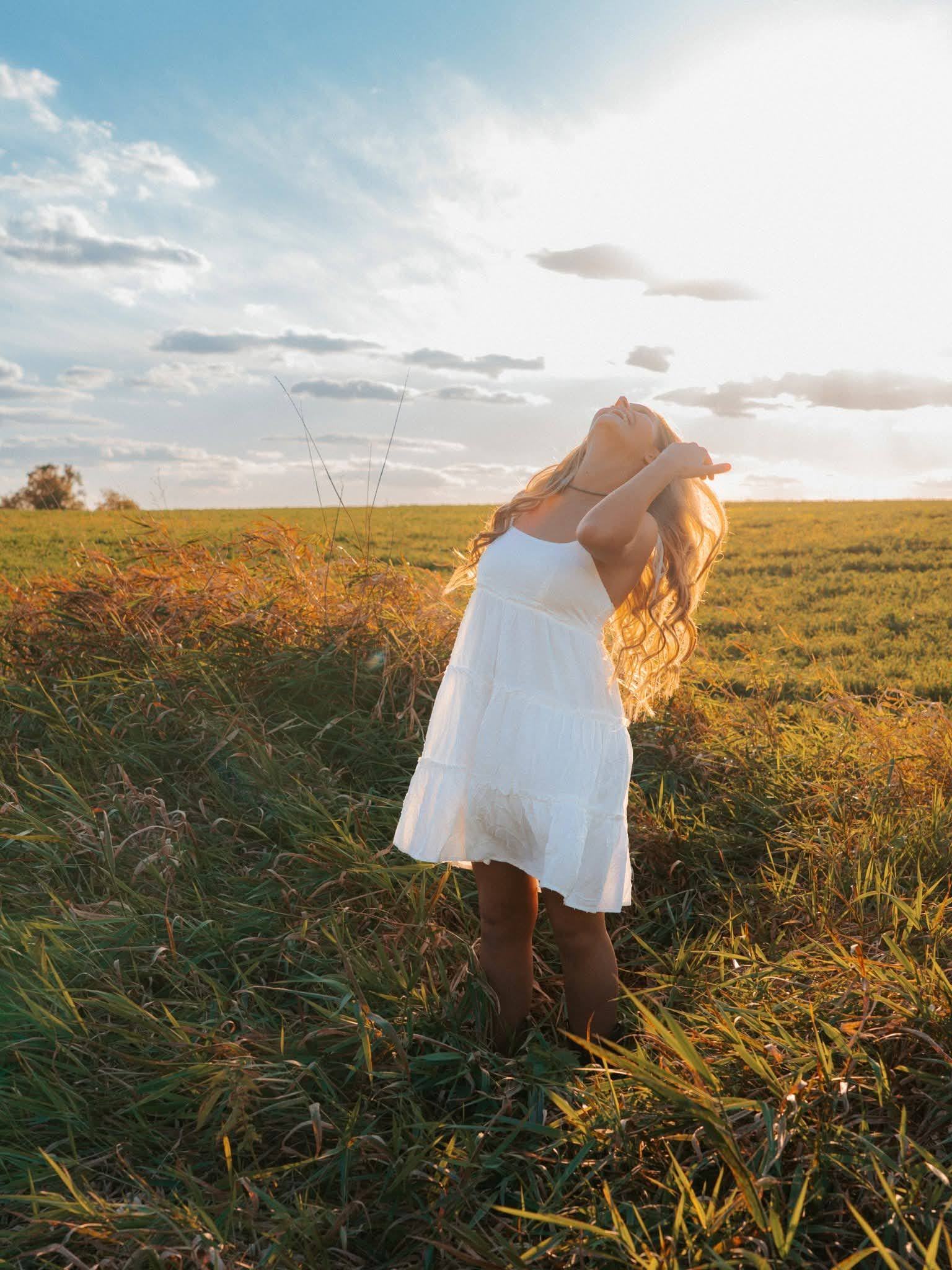 A senior in a white dress walking through tall prairie grass at sunset
