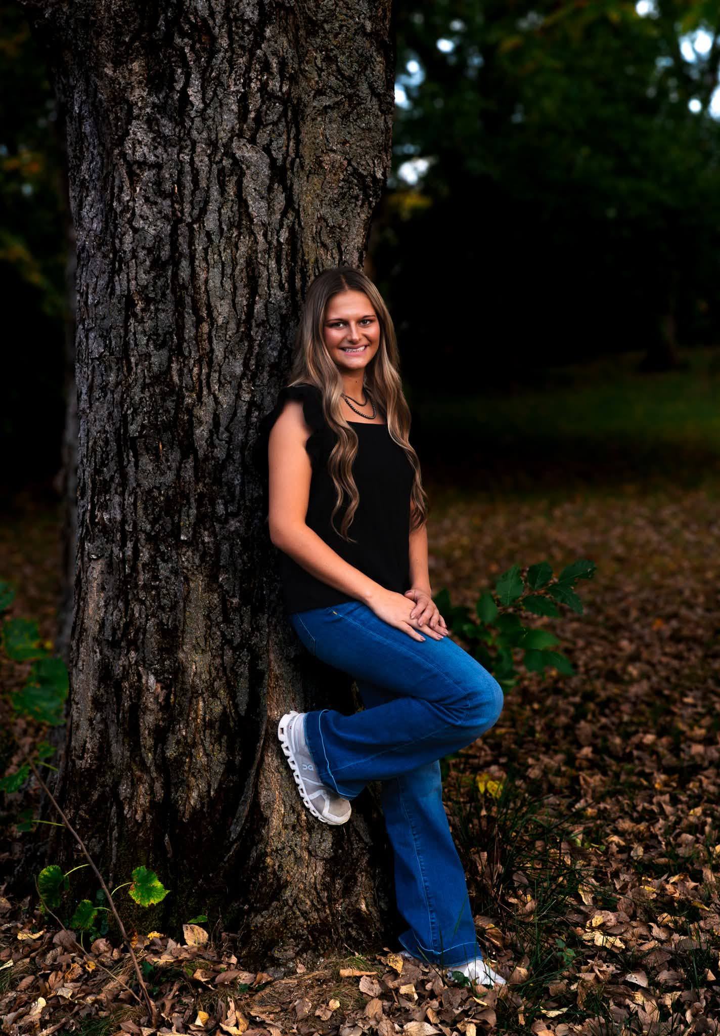 A senior leaning against a large tree trunk in a leaf-covered wood